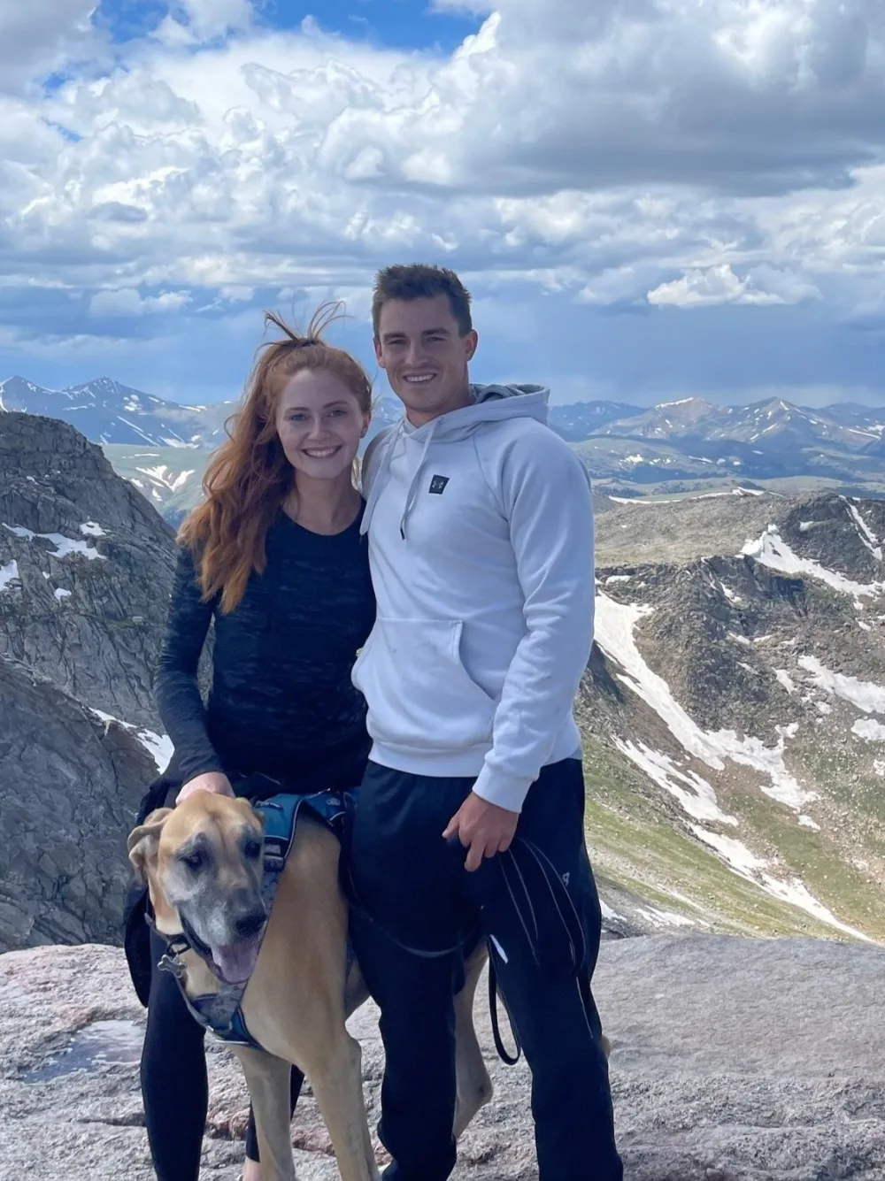 Couple with a large dog on a mountain summit, cloudy sky, snowy peaks in the background.